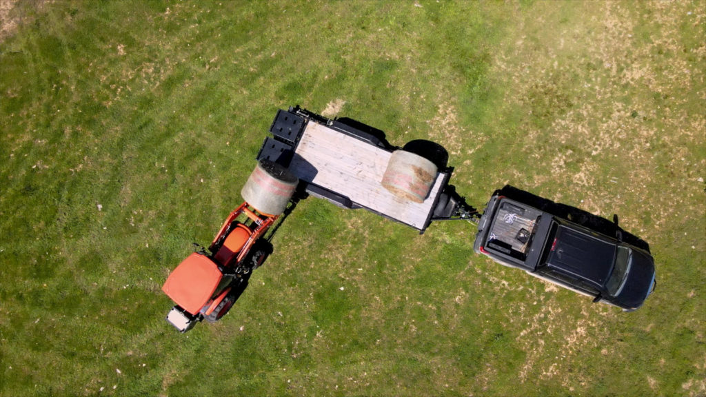 Tractor loading hay bale on to a trailer being pulled by a truck.